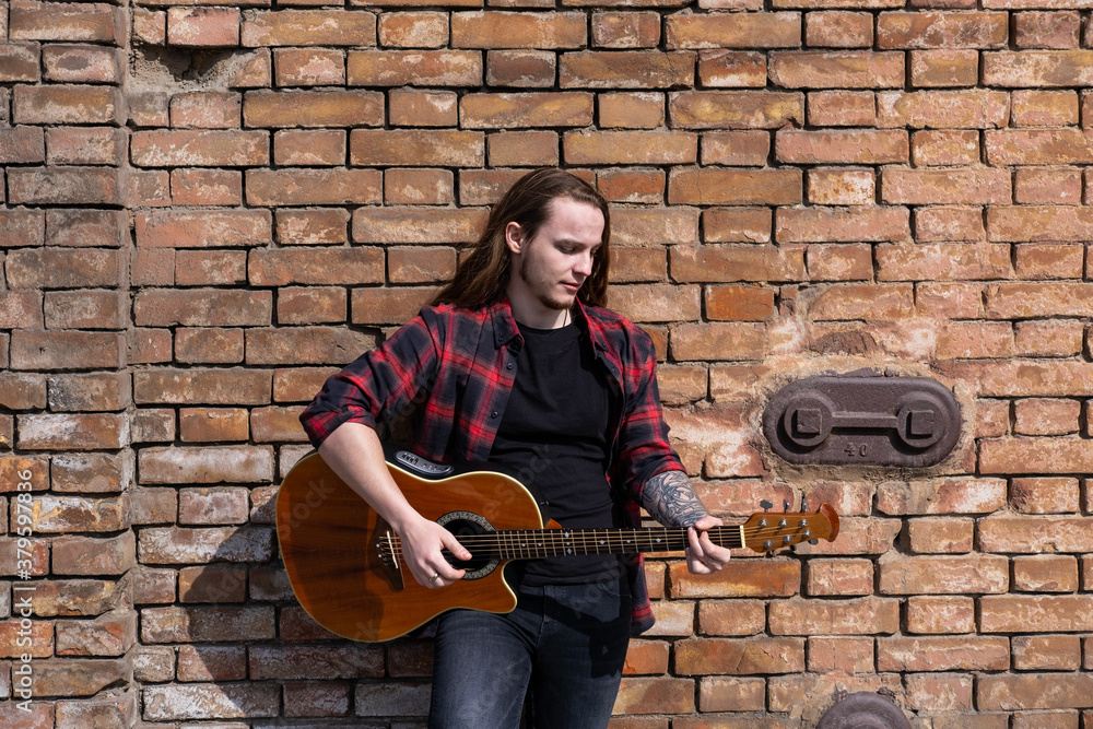 Young male with long hair and tattoo play on acoustic guitar outdoors on the street	