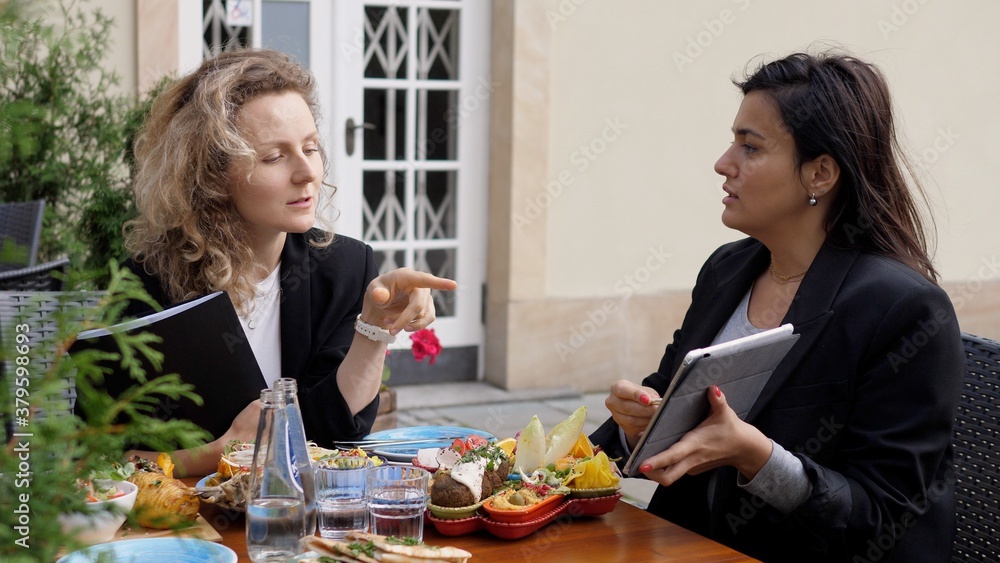 Brunette business woman showing graphs on a devise to her blonde colleague. Business meeting during lunch time Brunette business woman showing graphs on a devise to her blonde colleague. Business meeting during lunch time