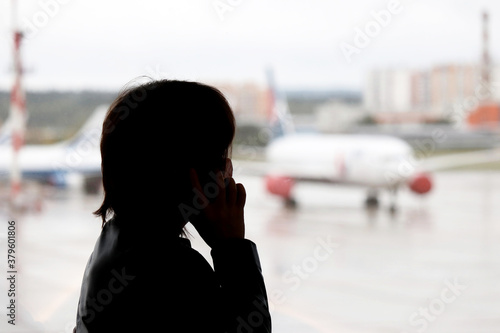 Silhouette of woman in mask talking on mobile phone in the airport terminal. Passenger waiting for their flight and looking to the airplanes through the window glass photo