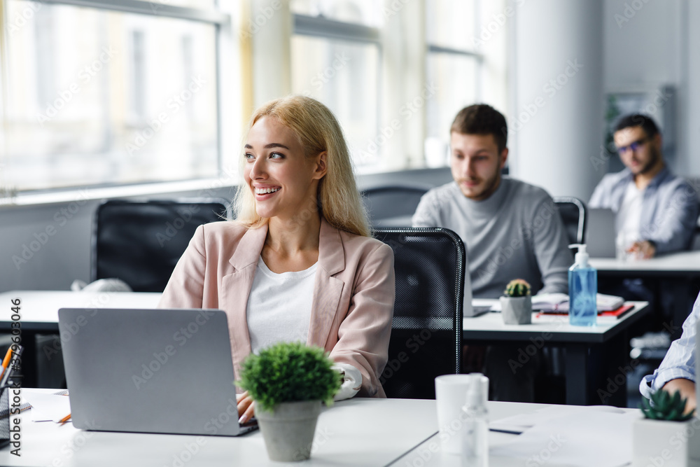 Work after lockdown. Happy young woman in suit looks out the window and works at laptop in office interior with plant in pot and antiseptics