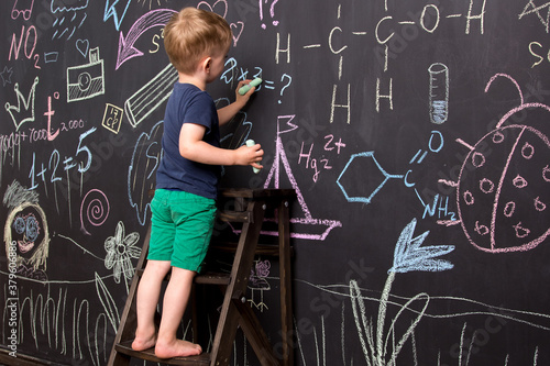 little boy draws with colorful chalk on a large blackboard. black wall with children's drawings and formulas. 