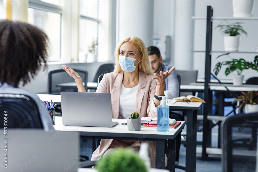 Conflict at work and problems with project. Angry woman in protective mask at workplace with laptop yelling at african american lady and gestures in interior of modern office Conflict at work and problems with project. Angry woman in protective mask at workplace with laptop yelling at african american lady and gestures in interior of modern office