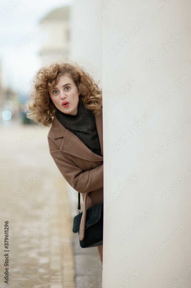A portrait of a curly-haired young woman peeking out from behind a white pillar. The girl looks in surprise, putting her lips in a bow