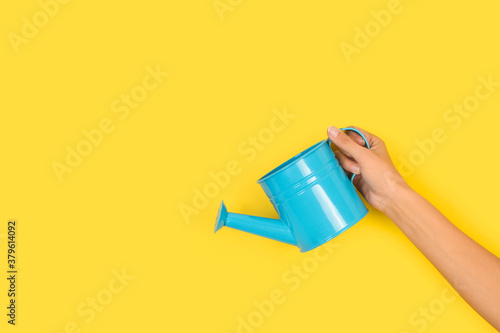 Mano de mujer sosteniendo una regadera azul sobre un fondo amarillo liso y aislado. Vista de frente. Copy space photo