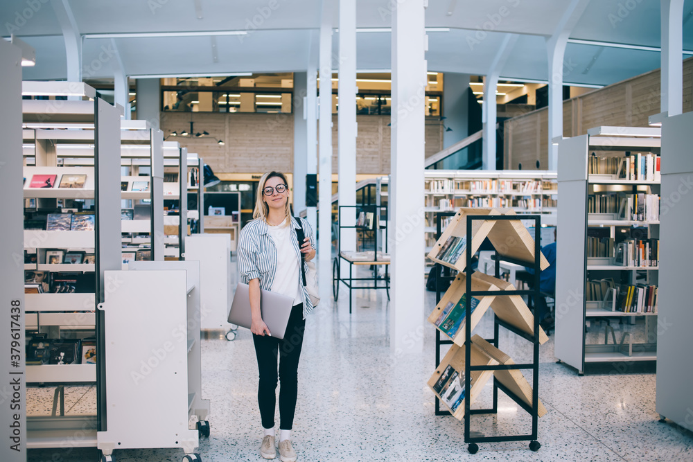 Full length portrait of young Caucasian journalist in classic spectacles and smart casual wear choosing place for remote job on modern laptop in coworking, beautiful woman with netbook posing indoors