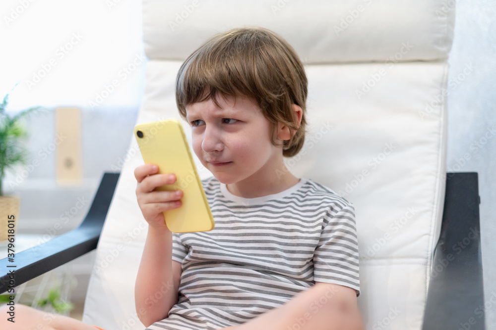 Offended little child boy playing online game, watching video on smartphone, sitting on couch entertaining in living room. Sad small kid using funny mobile apps, enjoying free leisure time at home