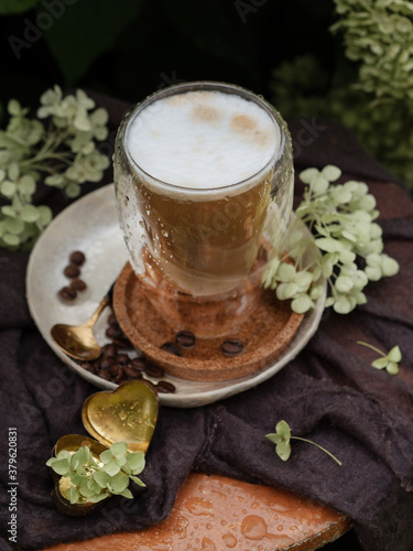 Cappuccino coffee is poured into a transparent glass with a double bottom. Coffee beans are poured nearby. Around there are hydrangea flowers and a vintage heart-shaped ornament. Breakfast in nature.