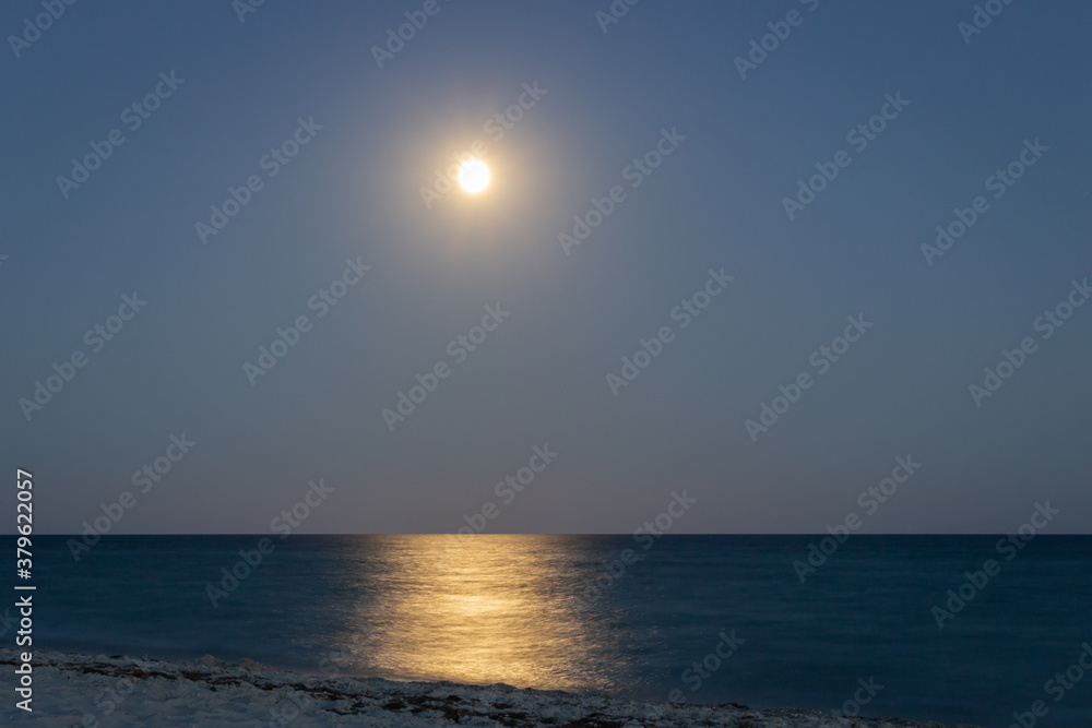 Fototapeta premium Full moon with moonlight on calm sea surface, long exposure. Island beach in the night. Moon path on sea. Twilight on seacoast. Scenic night sky over beach. Tranquility of summer night.