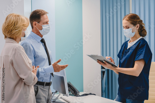 Caucasian middle aged couple having consultation and appointment in protective medical face masks at reception in clinic