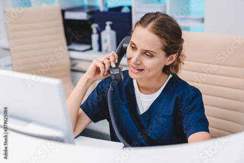 Young female receptionist talking on phone in clinic while sitting and looking on pc monitor
