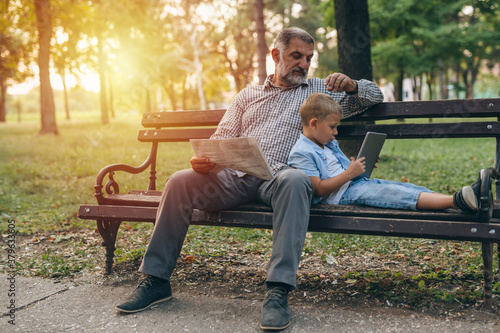 boy with his grandfather sitting on the bench and using tablet in the city park