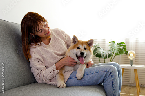 Young woman on grey textile sofa with nine months old japanese akita inu lying beside her. Female in denim pants and knitted sweater with funny big breed dog relaxing at home. Close up  copy space.