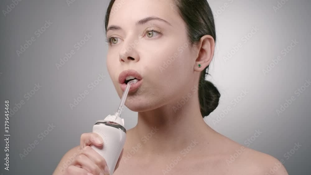 Young woman with dark hair ponytail cleaning her teeth with irrigator in bathroom. Morning healthcare routine with water flosser, mouthwashing. Light grey background. Slow motion 4k 100 fps Young woman with dark hair ponytail cleaning her teeth with irrigator in bathroom. Morning healthcare routine with water flosser, mouthwashing. Light grey background. Slow motion 4k 100 fps