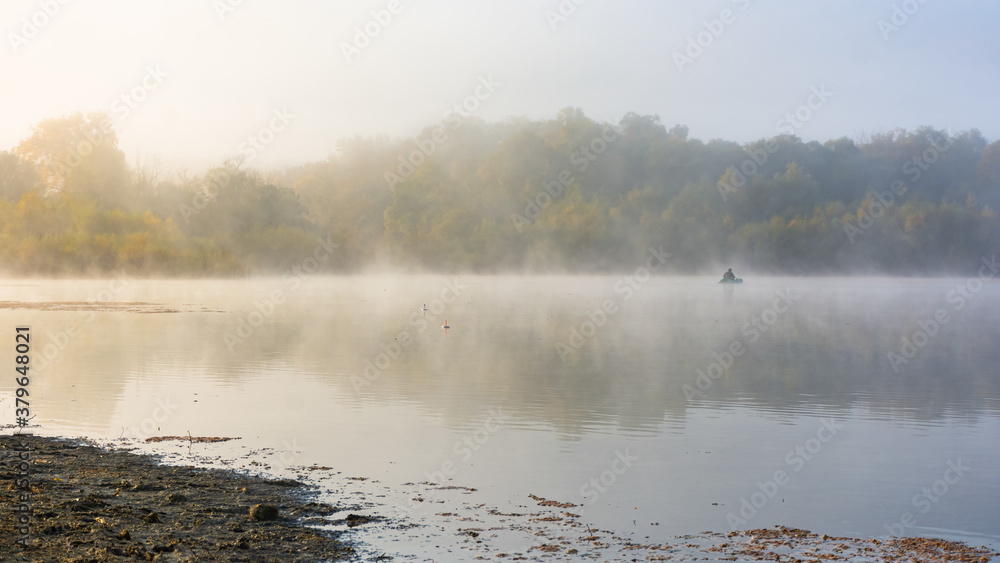 Autumn landscape with a fisherman on the lake in the fog. Early morning on the lake. A lone fisherman in a boat. A thick, gray morning fog. Relaxation in nature. Autumn landscape with a fisherman on the lake in the fog. Early morning on the lake. A lone fisherman in a boat. A thick, gray morning fog. Relaxation in nature.
