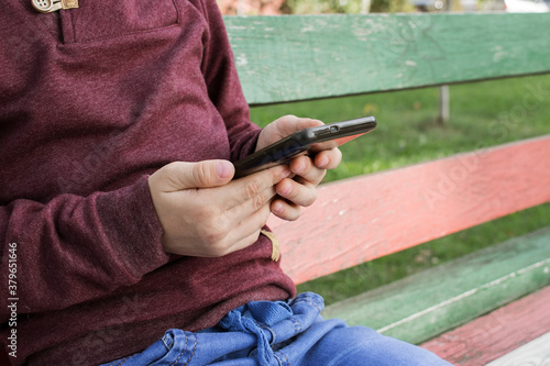 A boy holds a black phone in both hands while sitting on a Park bench. Maroon jacket and blue pants. Communication with my grandmother on the phone in nature