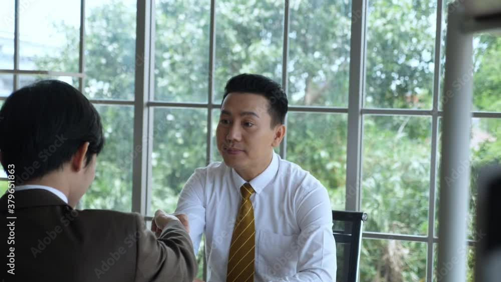 Business deal concept. Indoor, handheld, medium shot of Enthusiastic Asian businessman shaking hand, smiles, and talking to customer Business deal concept. Indoor, handheld, medium shot of Enthusiastic Asian businessman shaking hand, smiles, and talking to customer