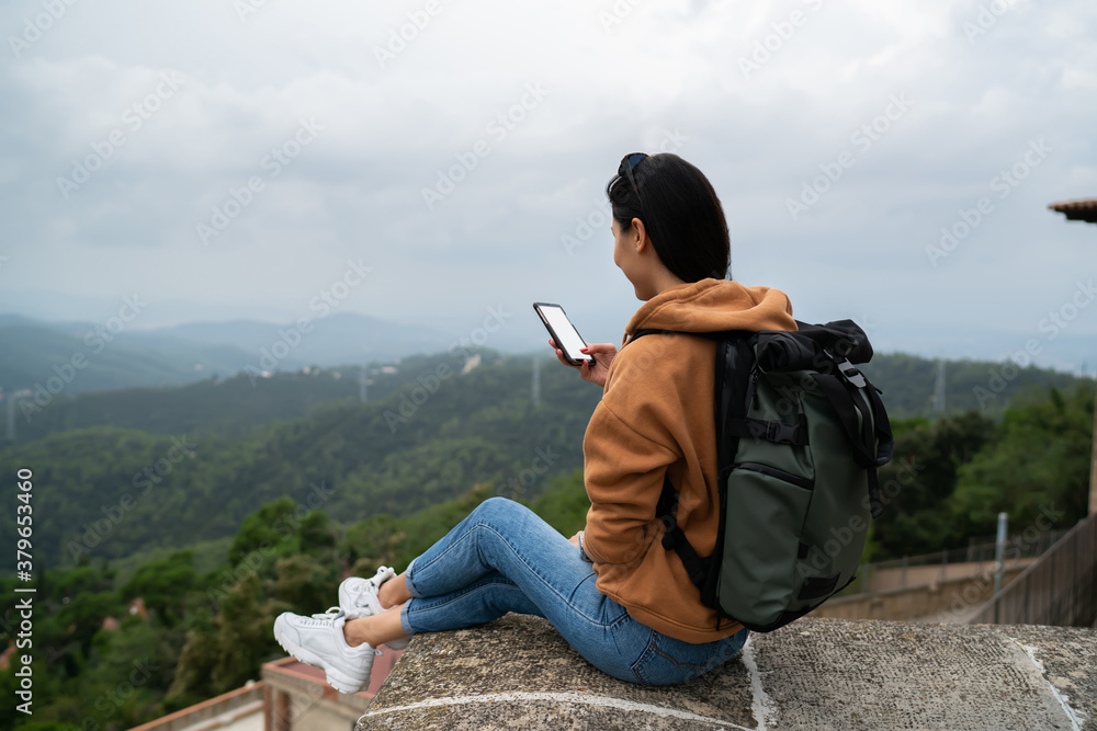 Back view of female traveler with backpack resting during destination holding smartphone with blank screen reading income mail, rear view of asian woman share content online via 4g on mobile phone