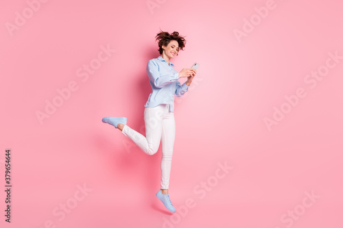 Full size profile photo of pretty lady good mood jump high up air carefree student browsing telephone chatting friends wear blue shirt white pants shoes isolated pink color background
