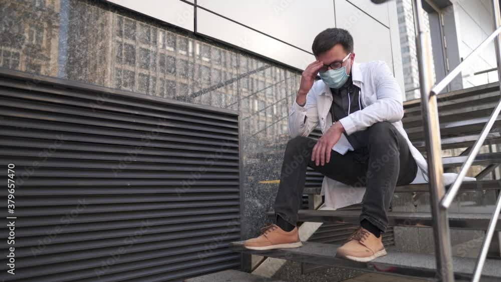 Caucasian young doctor man sits down on the stairs near the clinic building, tired and unhappy rubbing his nose and eyes, feeling tired and headache. Health care worker stress and frustration concept. Caucasian young doctor man sits down on the stairs near the clinic building, tired and unhappy rubbing his nose and eyes, feeling tired and headache. Health care worker stress and frustration concept.