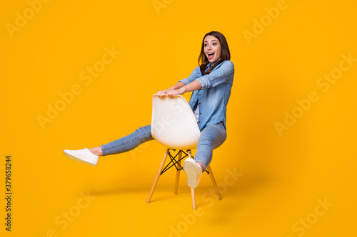Full body photo of attractive pretty funny lady good mood sitting chair spread legs hips fooling around playing wear casual denim shirt white shoes isolated bright yellow color background