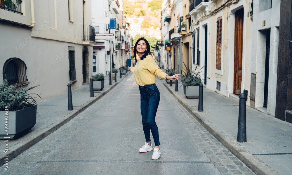 Full length portrait of funny tourist in headphones walking around touristic town dancing and smiling at camera, happy female generation Z in electronic equipment enjoying pastime vacations for travel