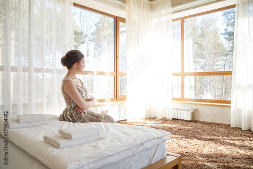 A beautiful girl bride in an elegant dress sits on a bed in a bright bedroom with large windows.