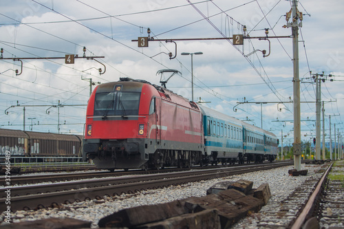 Modern electrical train  with red engine on track on a suburban station. Passenger train rushing towards the city in a suburban environment on a cloudy day. © Anze