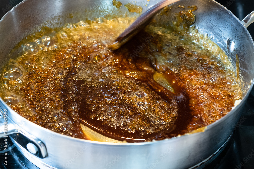 cooking salted butter caramel sauce at home kitchen - mixing boiled mixture of sugar and butter with wooden culinary spatula in steel pan close up cooking salted butter caramel sauce at home kitchen - mixing boiled mixture of sugar and butter with wooden culinary spatula in steel pan close up