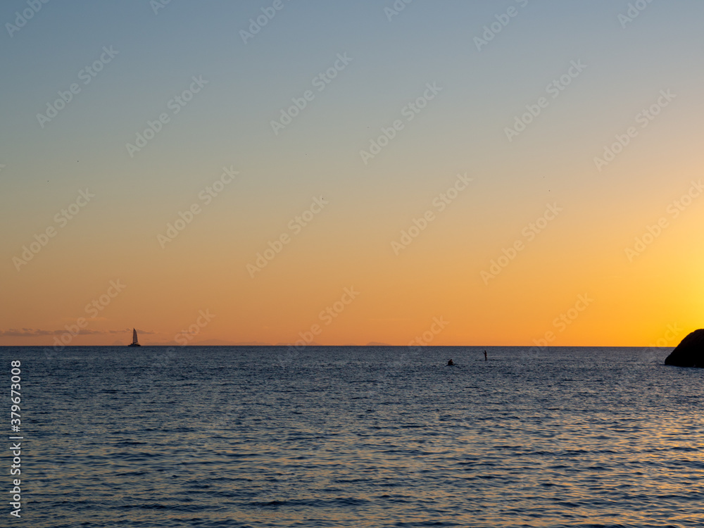 Beach in the Ibiza island with sailboat and yacht in the water during sunset. Boat and paddle surf silhouette in the ocean.