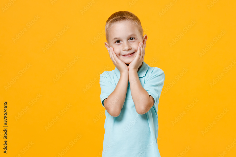 Smiling cute little fun male fair-haired brown-eyed kid boy 5-6 years old wearing stylish blue turquoise t-shirt polo put hands on face isolated on yellow color wall background, child studio portrait. Smiling cute little fun male fair-haired brown-eyed kid boy 5-6 years old wearing stylish blue turquoise t-shirt polo put hands on face isolated on yellow color wall background, child studio portrait.