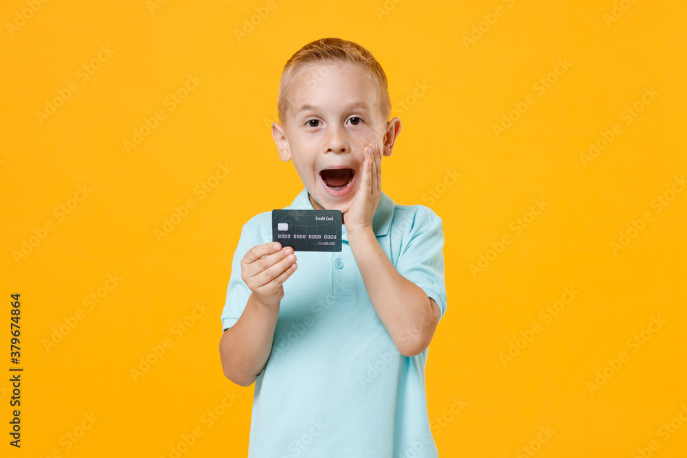 Excited happy little fun male kid boy 5-6 years old wearing stylish blue turquoise t-shirt polo hold in hand mockups of credit bank card isolated on yellow color wall background child studio portrait. Excited happy little fun male kid boy 5-6 years old wearing stylish blue turquoise t-shirt polo hold in hand mockups of credit bank card isolated on yellow color wall background child studio portrait.