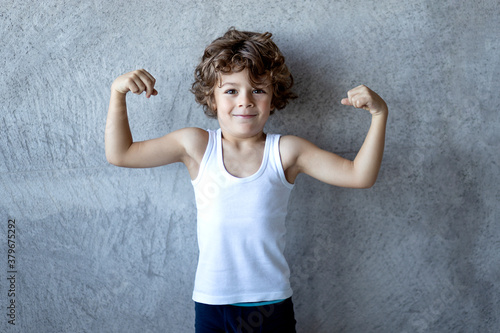 Принти на полотні Little curly haired red boy in holding fists up and showing biceps on concrete grey background