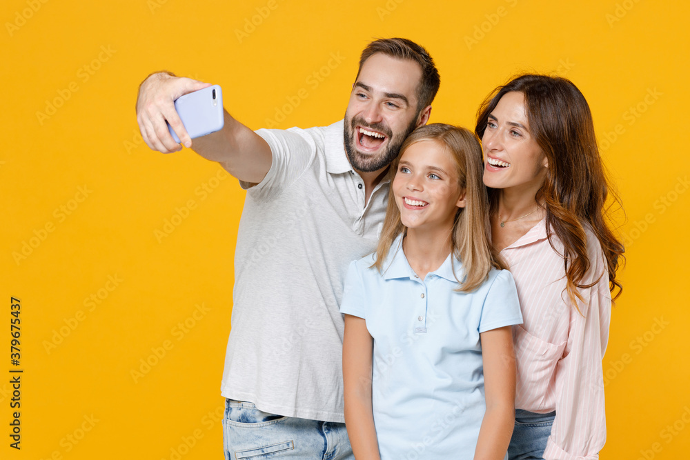 Cheerful laughing funny young parents mom dad with child kid daughter teen girl in basic t-shirts doing selfie shot on mobile phone isolated on yellow background studio portrait. Family day concept. Cheerful laughing funny young parents mom dad with child kid daughter teen girl in basic t-shirts doing selfie shot on mobile phone isolated on yellow background studio portrait. Family day concept.