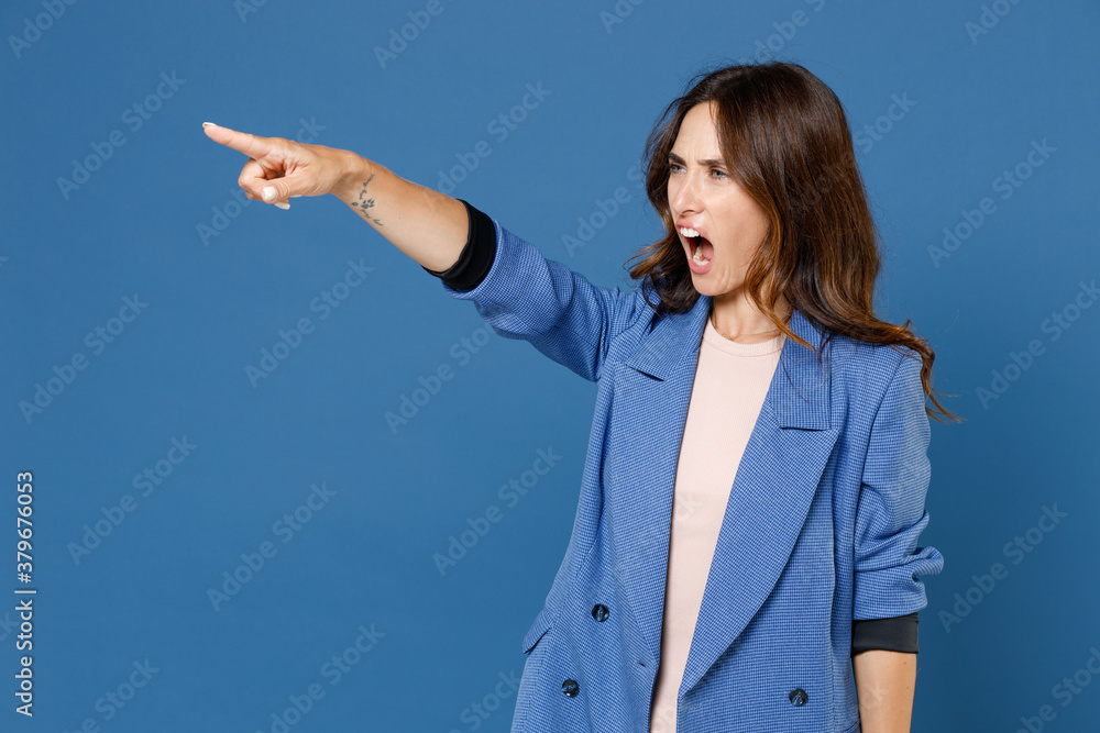 Angry irritated displeased dissatisfied young brunette woman 20s wearing basic jacket standing pointing index finger aside screaming swearing isolated on bright blue colour background studio portrait. Angry irritated displeased dissatisfied young brunette woman 20s wearing basic jacket standing pointing index finger aside screaming swearing isolated on bright blue colour background studio portrait.