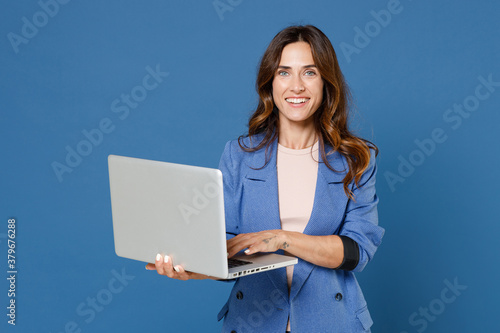 Smiling cheerful beautiful attractive young brunette woman 20s wearing basic jacket standing working on laptop pc computer looking camera isolated on bright blue colour background studio portrait.