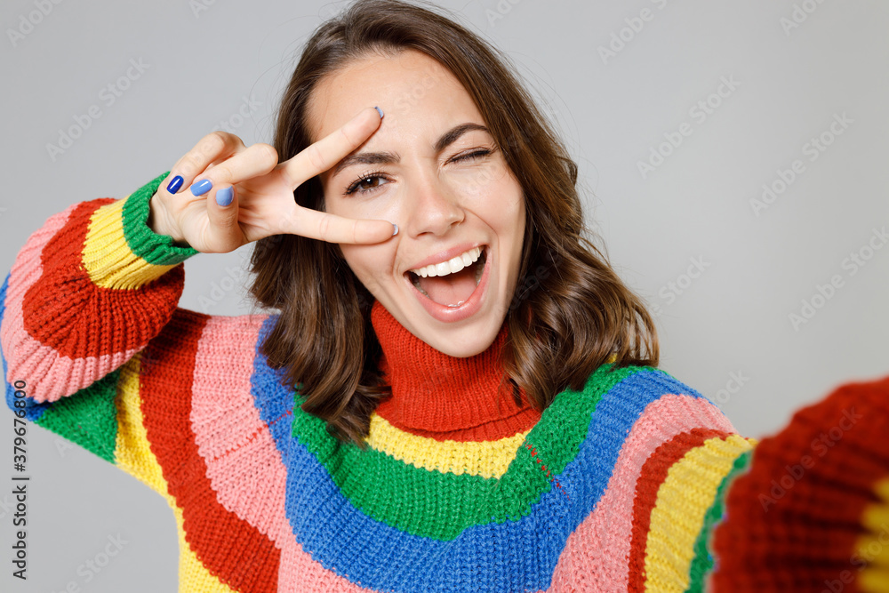 Close up of funny blinking young woman 20s wearing casual colorful knitted sweater standing doing selfie shot on mobile phone showing victory sign isolated on grey colour background, studio portrait. Close up of funny blinking young woman 20s wearing casual colorful knitted sweater standing doing selfie shot on mobile phone showing victory sign isolated on grey colour background, studio portrait.