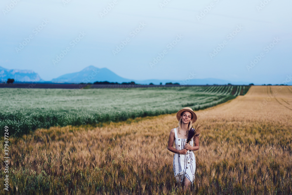 Half length portrait of happy woman with flowers bouquet posing in barley field and smiling at camera during solo journey to rural countryside, pretty female with posy discovering nature environment