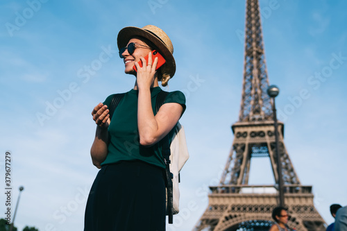 Smiling female tourist in trendy apparel enjoying cellphone conversation in roaming for discussing solo vacations in France, happy woman in sunglasses laughing during smartphone calling via app