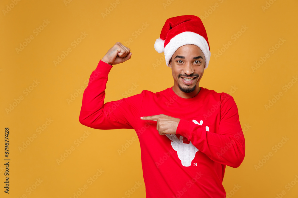 Smiling young Santa african american man in red sweater Christmas hat pointing index finger on biceps muscles isolated on yellow background studio. Happy New Year celebration merry holiday concept.