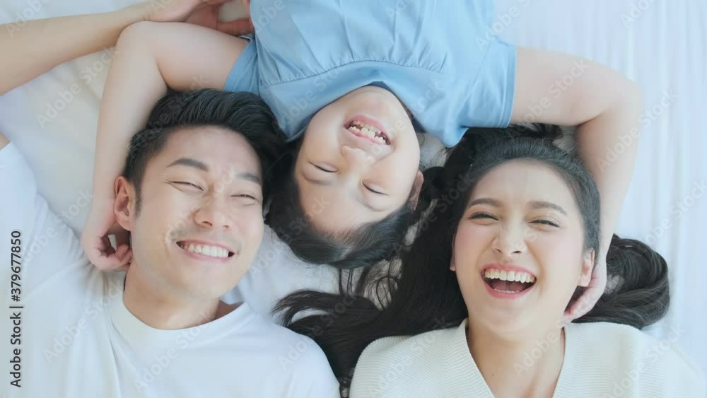 Happy Asian family mom dad and daughter child laying laugh smile on white soft bed in bedroom relax casual leisure weekend activity, top view handheld shot Happy Asian family mom dad and daughter child laying laugh smile on white soft bed in bedroom relax casual leisure weekend activity, top view handheld shot