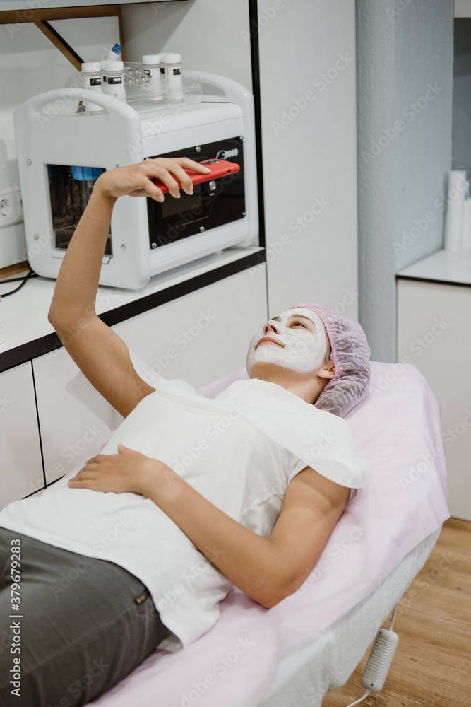 Professional skin care in beauty salon. Young woman with clay face mask takes selfie on phone. Facial care by beautician at spa salon. Acne Treatment, face peeling mask, spa beauty treatment