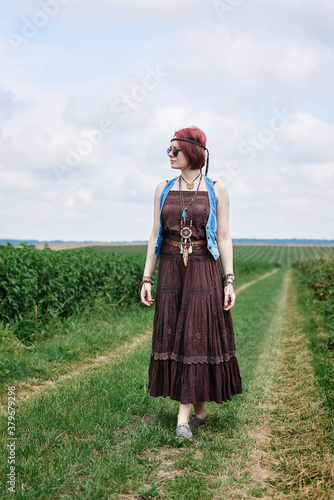 Young hippie woman with short red hair, wearing boho style clothes and sunglasses, standing on green currant field, posing for picture. Female portrait on natural background. Eco tourism concept.