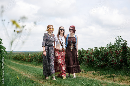 Three young hippie women, wearing boho style clothes, walking on green currant field in summer. Eco tourism concept. Friends traveling in rural countryside.