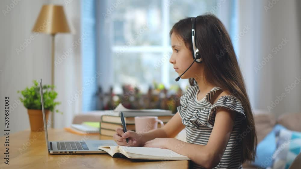 Positive kid in headset writing on notebook and using laptop at home studying online Positive kid in headset writing on notebook and using laptop at home studying online