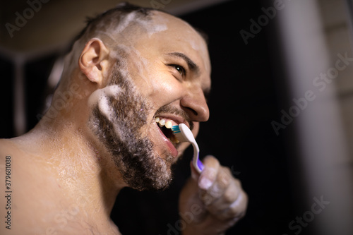 Young smiling handsome man in foam brushing teeth with toothbrush while taking shower, side view, close up. Lifestyle, Morning routine, oral hygiene, personal care, water saving concept