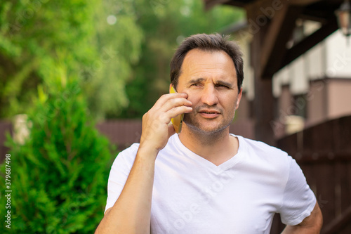 Closeup portrait view of one handsome unshaven squinting 40s man in white T-shirt speaking on mobile phone outdoor on blurred green natural background, horizontal picture. concept of equanimity