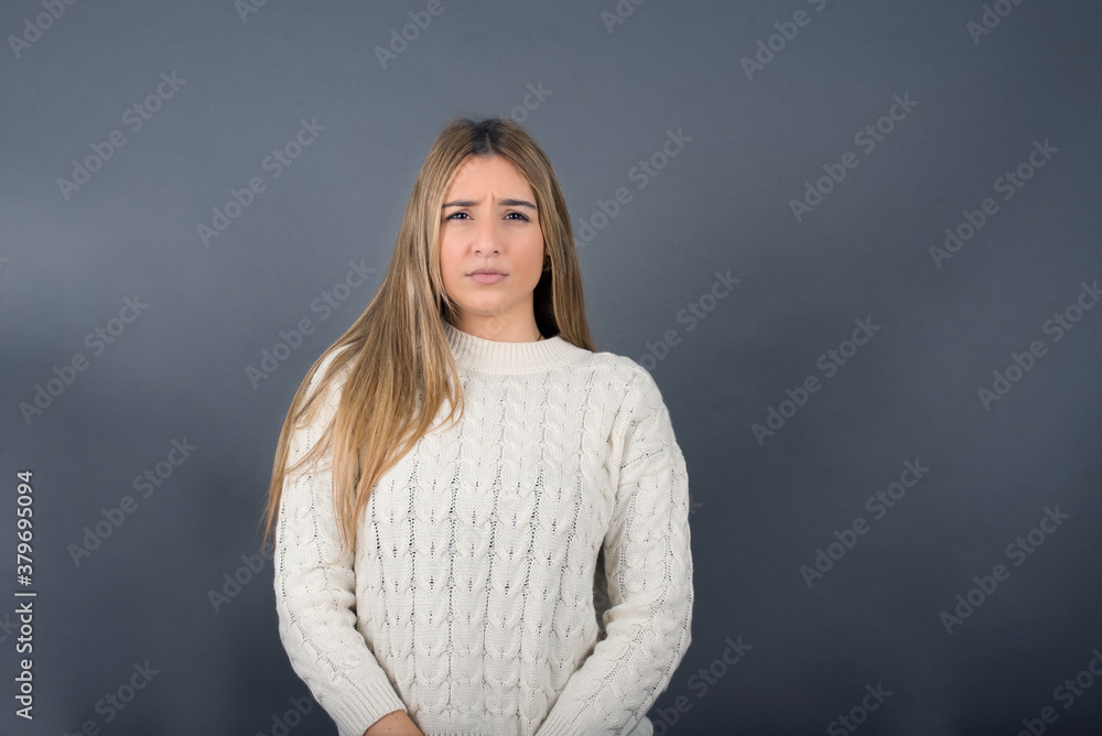 Beautiful blonde young woman wearing white sweater feeling confident against gray studio background. Beautiful blonde young woman wearing white sweater feeling confident against gray studio background.