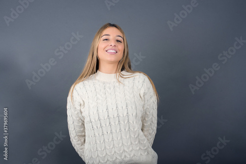 Beautiful blonde young woman wearing white sweater feeling confident against gray studio background.