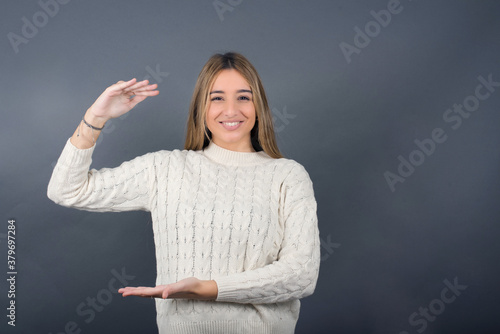 Beautiful young blonde woman wearing white sweater standing against gray background, 