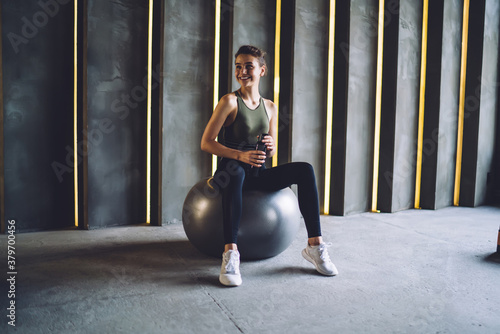 Smiling beautiful brunette young woman sitting on fitball resting after resultative workout keeping healthy lifestyle, caucasian woman fitness instructor enjoying break and refreshment in gym